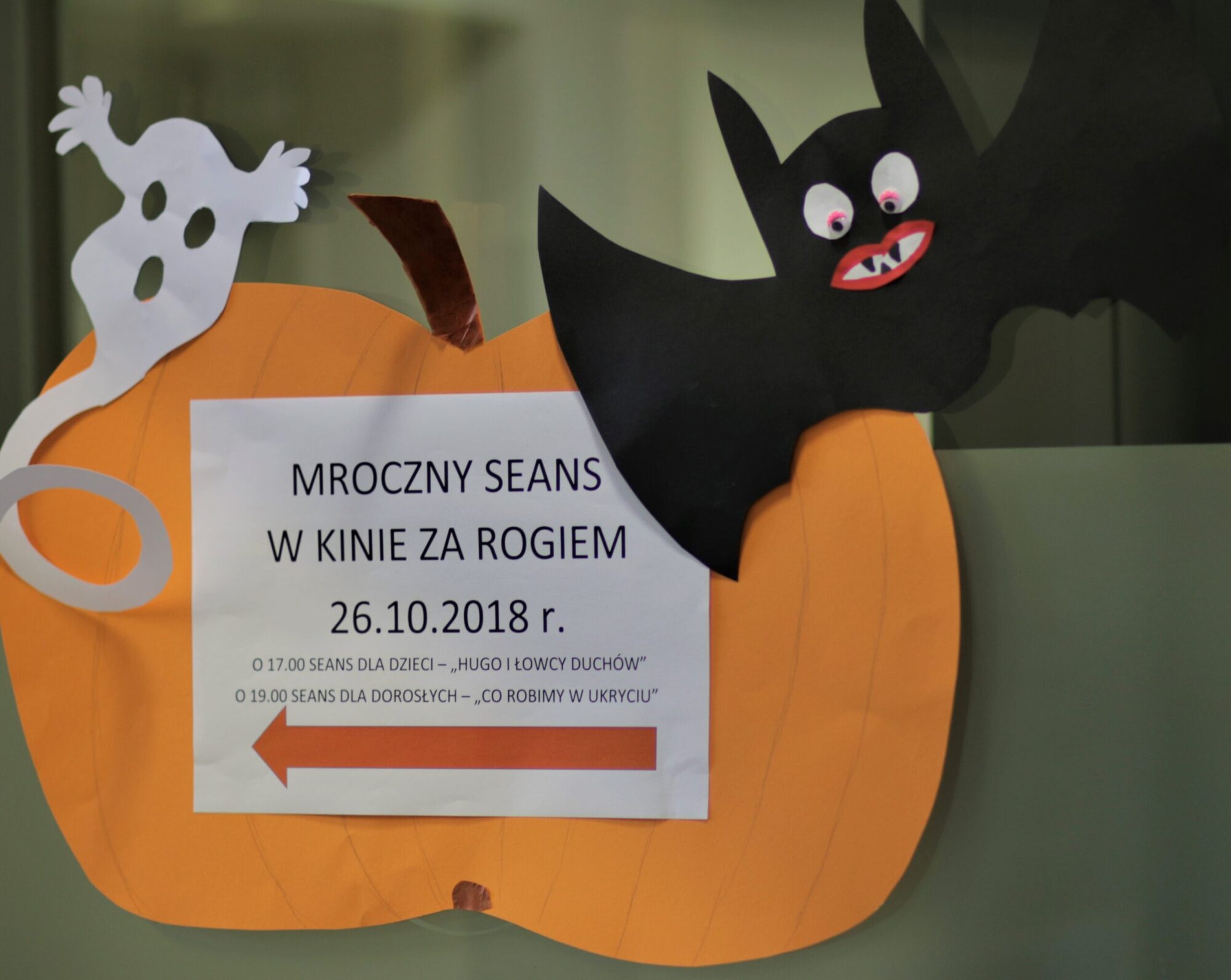 Znak o tematyce Halloween z wyciętymi z papieru białym duchem, czarnym nietoperzem i pomarańczową dynią. Znak reklamuje upiorny wieczór filmowy 26 października 2018 r. z godzinami seansów dla dzieci i dorosłych.