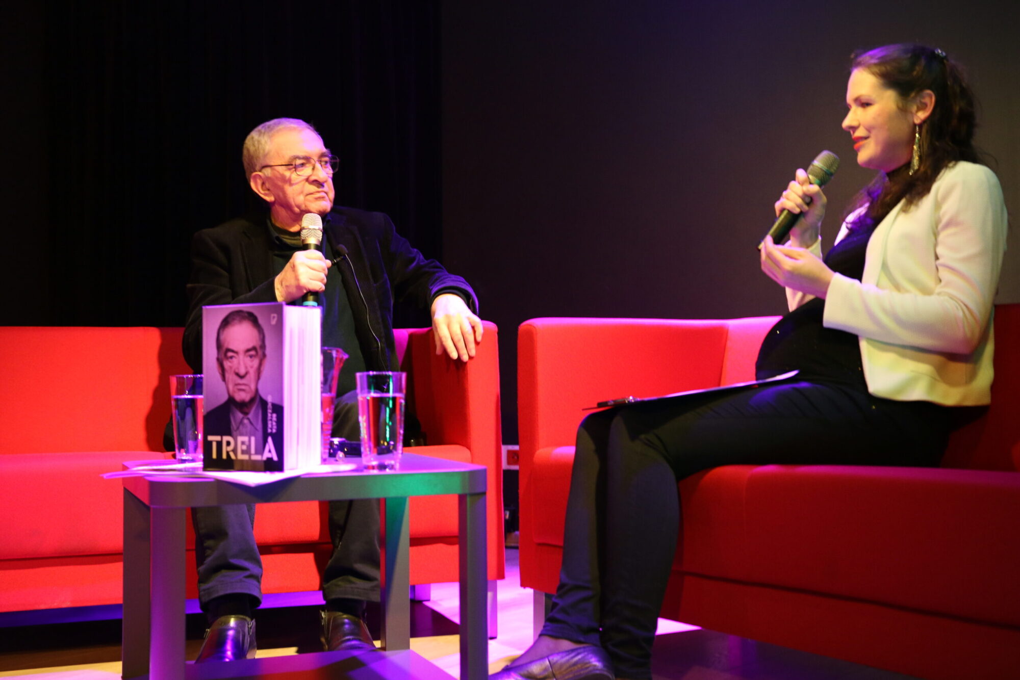 Mężczyzna i kobieta siedzą na czerwonych kanapach, trzymają mikrofony i rozmawiają. Na stole między nimi leży książka z portretem mężczyzny i tytułem TRELA. Na stole znajdują się również szklanki z wodą.