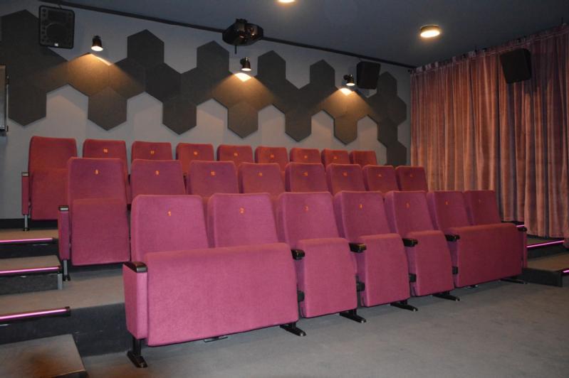 Małe kino z trzema rzędami różowych tapicerowanych siedzeń, szarymi geometrycznymi panelami ściennymi i różowymi zasłonami zasłaniającymi jedną ścianę. Kino jest puste i delikatnie oświetlone.