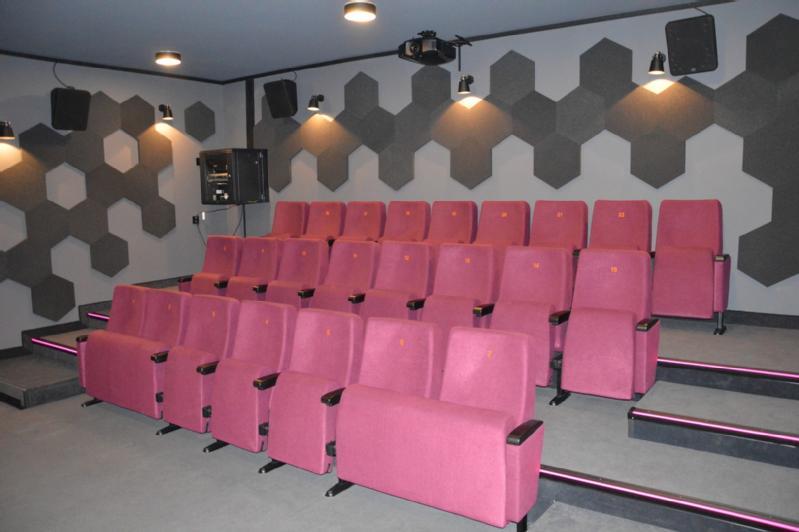 Mała nowoczesna sala kinowa z trzema rzędami różowych, wyściełanych siedzeń, szarymi sześciokątnymi panelami ściennymi do akustyki oraz głośnikami i projektorem zamontowanymi na suficie. Sala ma szarą wykładzinę i słabe oświetlenie sufitowe.