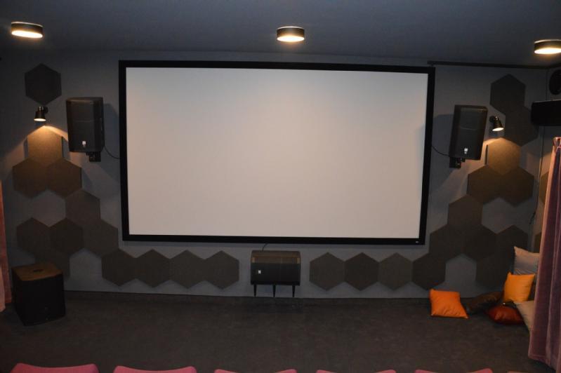Niewielka sala kinowa z dużym, pustym ekranem, sześciokątnymi panelami akustycznymi na ścianach, głośnikami i kilkoma kolorowymi poduszkami na podłodze przed ekranem.