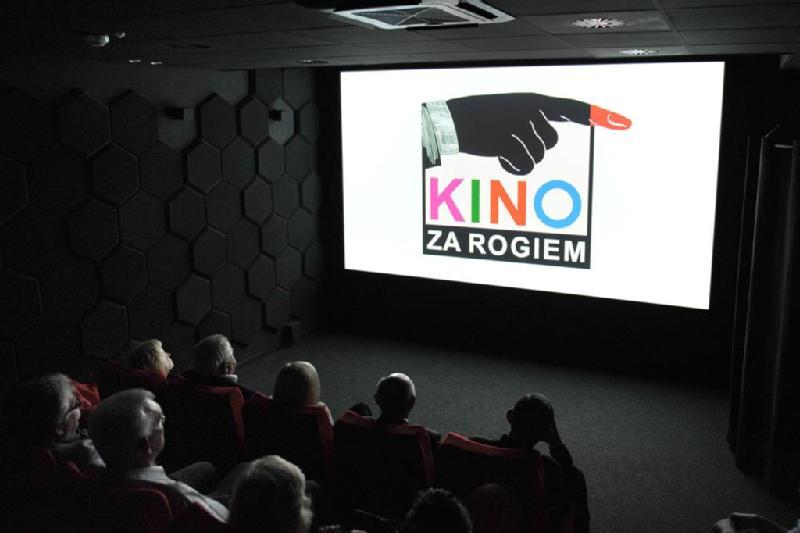 Niewielka grupa ludzi siedzi w ciemnym kinie, patrząc na ekran wyświetlający logo z wskazującą ręką i napis Kino Za Rogiem w kolorowych literach.