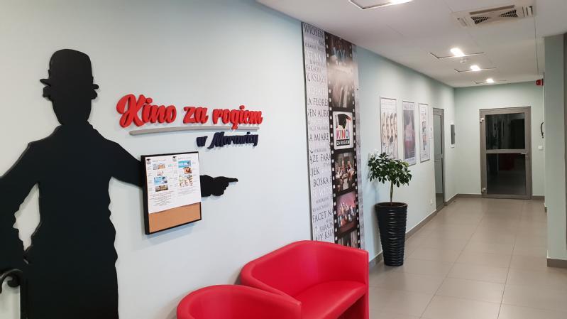 Korytarz z plakatami filmowymi na ścianie, czerwonym fotelem, rośliną doniczkową i sylwetką mężczyzny w kapeluszu. Na lewej ścianie wisi tabliczka z napisem Kino za Rogiem w Morawicy.