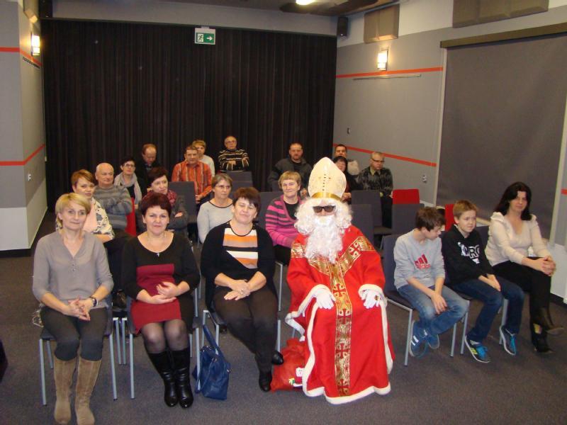 Grupa dorosłych i dzieci siedzi w środku, pozując do zdjęcia. W pierwszym rzędzie siedzi osoba przebrana za Świętego Mikołaja w czerwonej szacie i mitrze. Atmosfera wydaje się świąteczna.