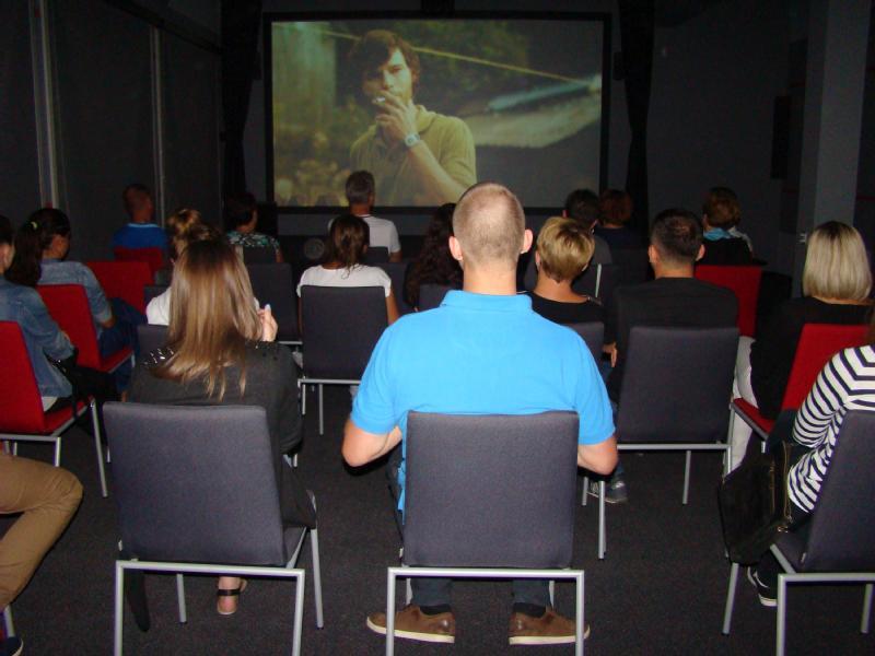 Ludzie siedzą w rzędach krzeseł w małym, ciemnym pokoju, oglądając film wyświetlany na ekranie z przodu. Większość widzów patrzy przed siebie, a w pokoju panuje swobodna, intymna atmosfera.