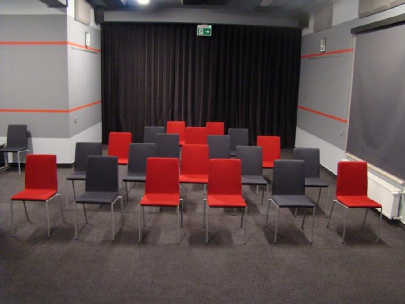 Mały pokój z 16 krzesłami ustawionymi w czterech rzędach. Krzesła są naprzemiennie w kolorze czerwonym i szarym. Z przodu jest czarna zasłona, a ściany są szare z czerwonymi paskami.