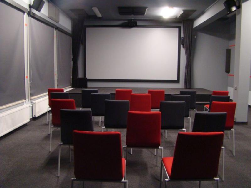 Mała, pusta sala projekcyjna z białym ekranem projekcyjnym, szarymi ścianami i rzędami czarnych i czerwonych krzeseł zwróconych w stronę ekranu. Sala ma minimalne oświetlenie i zamknięte rolety okienne.