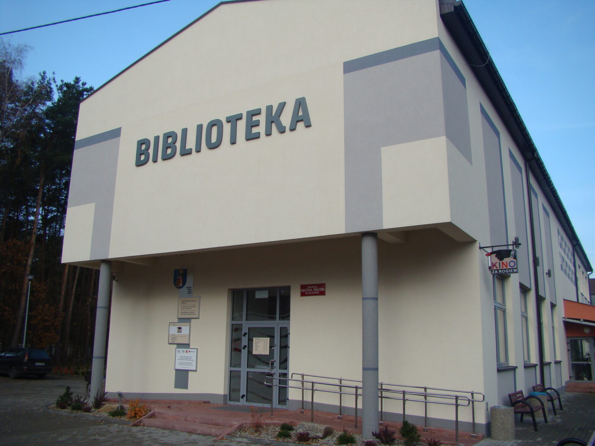 Nowoczesny, jasny budynek z napisem BIBLIOTEKA widocznym nad wejściem, wskazującym, że jest to biblioteka. Z boku znajduje się podjazd dla wózków inwalidzkich i mały znak kina.