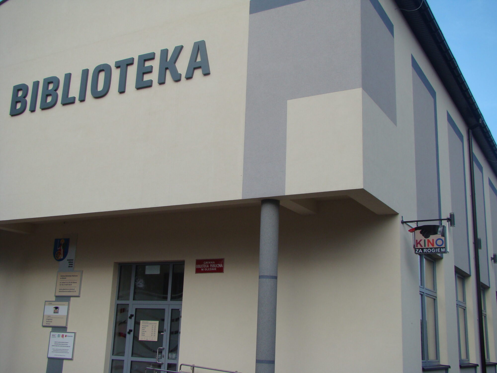 Nowoczesny budynek z napisem BIBLIOTEKA na fasadzie, wskazującym na bibliotekę. Są drzwi, kilka tabliczek i mały znak z napisem KINO ZA ROGIEM przy wejściu.