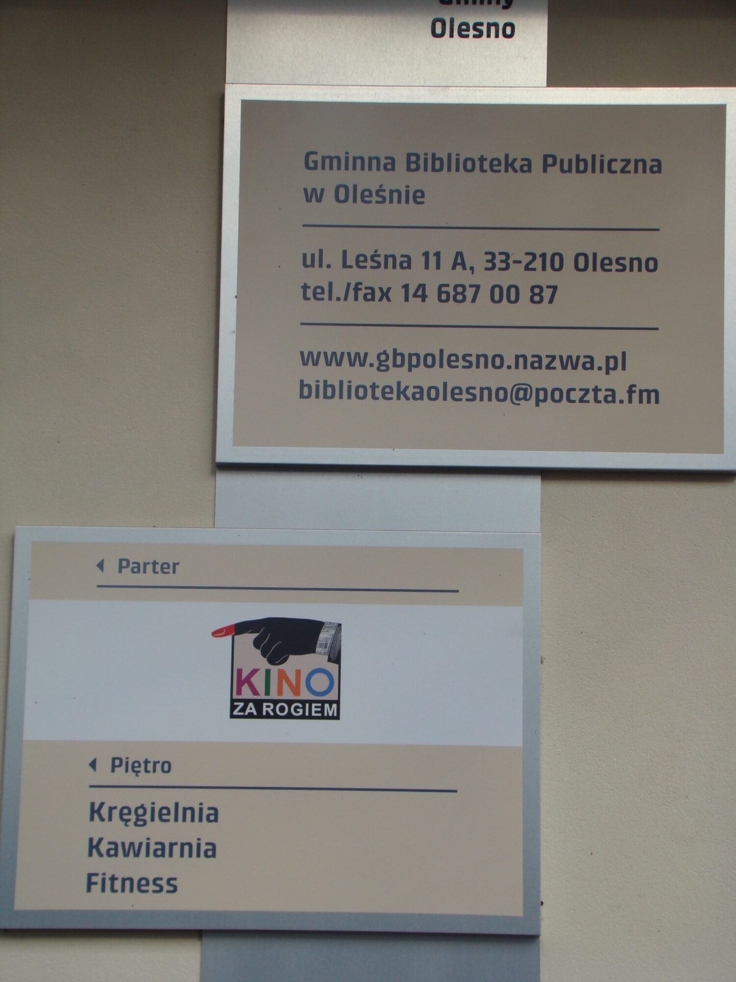 Dwa znaki na ścianie w języku polskim. Górny znak wyświetla dane kontaktowe Biblioteki Publicznej w Oleśnie, w tym adres, telefon, stronę internetową i e-mail. Dolny znak zawiera listę kin, kręgielni, kawiarni i siłowni.