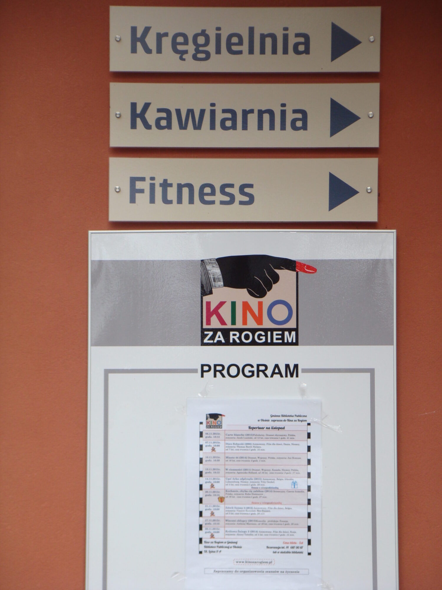 Trzy znaki kierunkowe w języku polskim wskazują na kręgielnię, kawiarnię i centrum fitness. Poniżej plakat przedstawia program Kino za Rogiem z ręką wskazującą na logo kina.