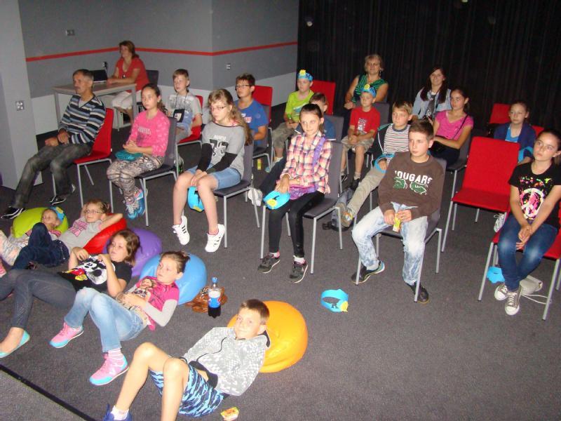 Grupa dzieci i kilku dorosłych siedzi na krzesłach i kolorowych pufach w pokoju, patrząc przed siebie i oglądając coś, prawdopodobnie film lub prezentację. Atmosfera wydaje się swobodna i zrelaksowana.