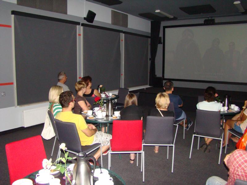 Grupa ludzi siedzi na krzesłach w słabo oświetlonym pokoju, oglądając film wyświetlany na dużym ekranie. Na pierwszym planie znajdują się stoły z kubkami, talerzami i kawiarką. Okna są zasłonięte żaluzjami.