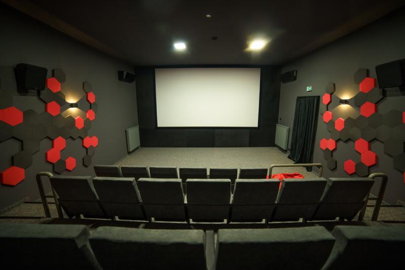 Małe nowoczesne kino z szarymi siedzeniami, geometrycznymi sześciokątnymi dekoracjami ściennymi w kolorze czarnym i czerwonym oraz pustym białym ekranem z przodu. Sala jest pusta, z wyjątkiem jednej czerwonej kurtki przewieszonej przez siedzenie.