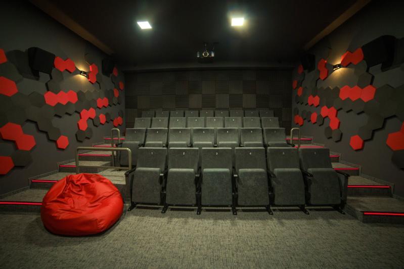 Małe, nowoczesne kino domowe z szarymi, piętrowymi siedzeniami, pojedynczym czerwonym fotelem-workiem z przodu i ścianami ozdobionymi czarnymi i czerwonymi heksagonalnymi panelami akustycznymi. Nad nimi widoczne są lampy sufitowe i projektor.