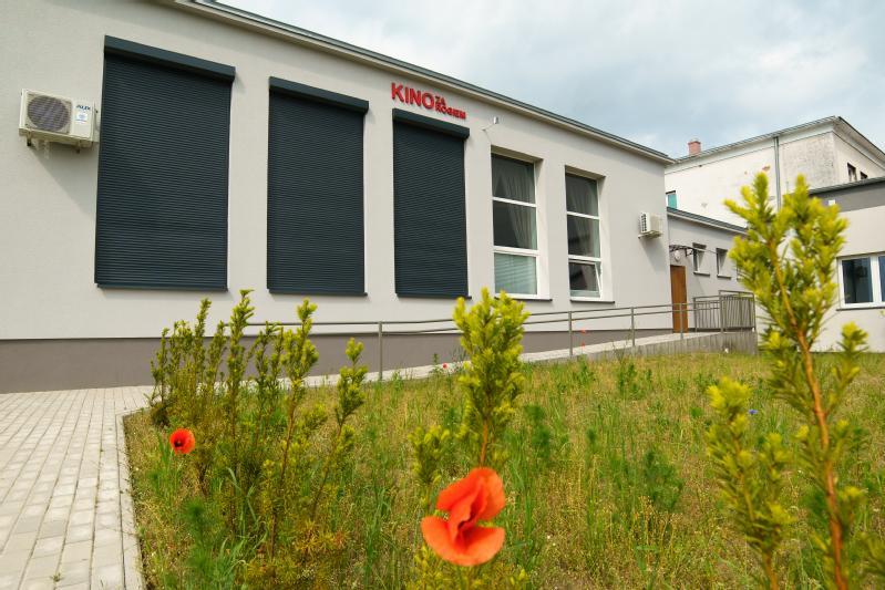 Nowoczesny, parterowy budynek kina z napisem KINO napisanym czerwonym kolorem nad oknami, czarnymi okiennicami i rampą, otoczony trawiastym obszarem z czerwonymi kwiatami maku na pierwszym planie.