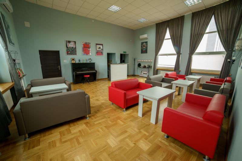 Nowoczesny salon z czerwonymi i szarymi fotelami, białymi stolikami, pianinem przy ścianie, dużymi oknami z zasłonami i oprawionymi obrazami na jasnoniebieskich ścianach. Pokój ma drewnianą podłogę i wysoki sufit.