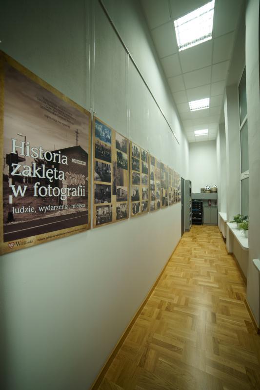 Korytarz z drewnianymi podłogami i wysokimi oknami ma wystawę zdjęć na lewej ścianie. Wystawa obejmuje duży plakat w języku polskim i kilka historycznych fotografii ułożonych w rzędach.