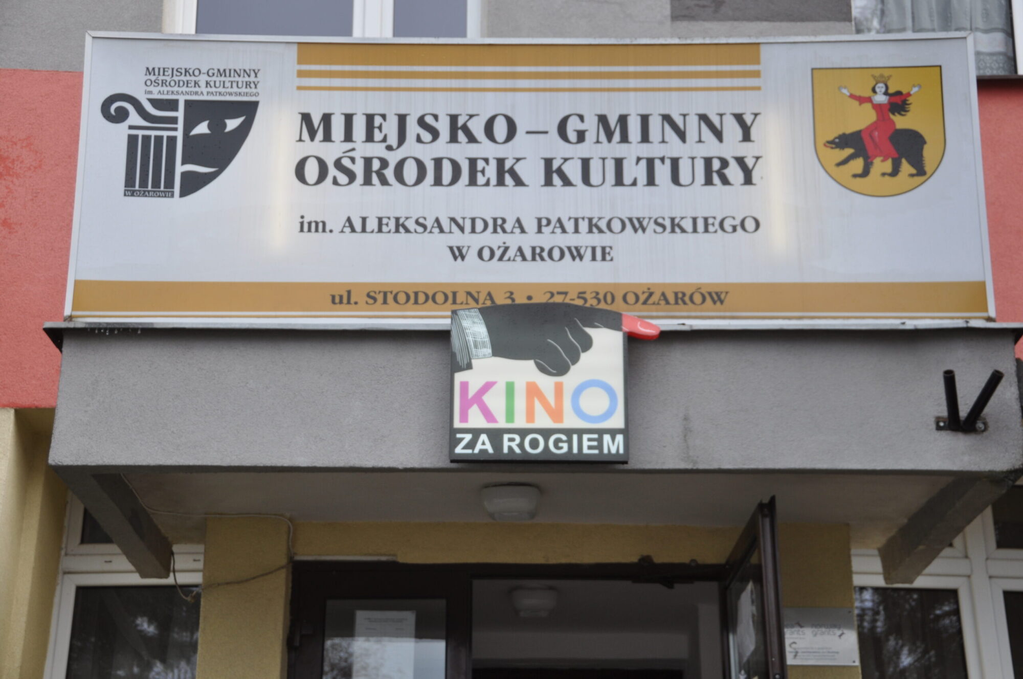 Wejście do budynku, nad którym widnieją dwa szyldy: jeden z nich to Miejsko-Gminny Ośrodek Kultury z herbem, a pod nim znajduje się mniejszy kolorowy szyld z napisem Kino za Rogiem.