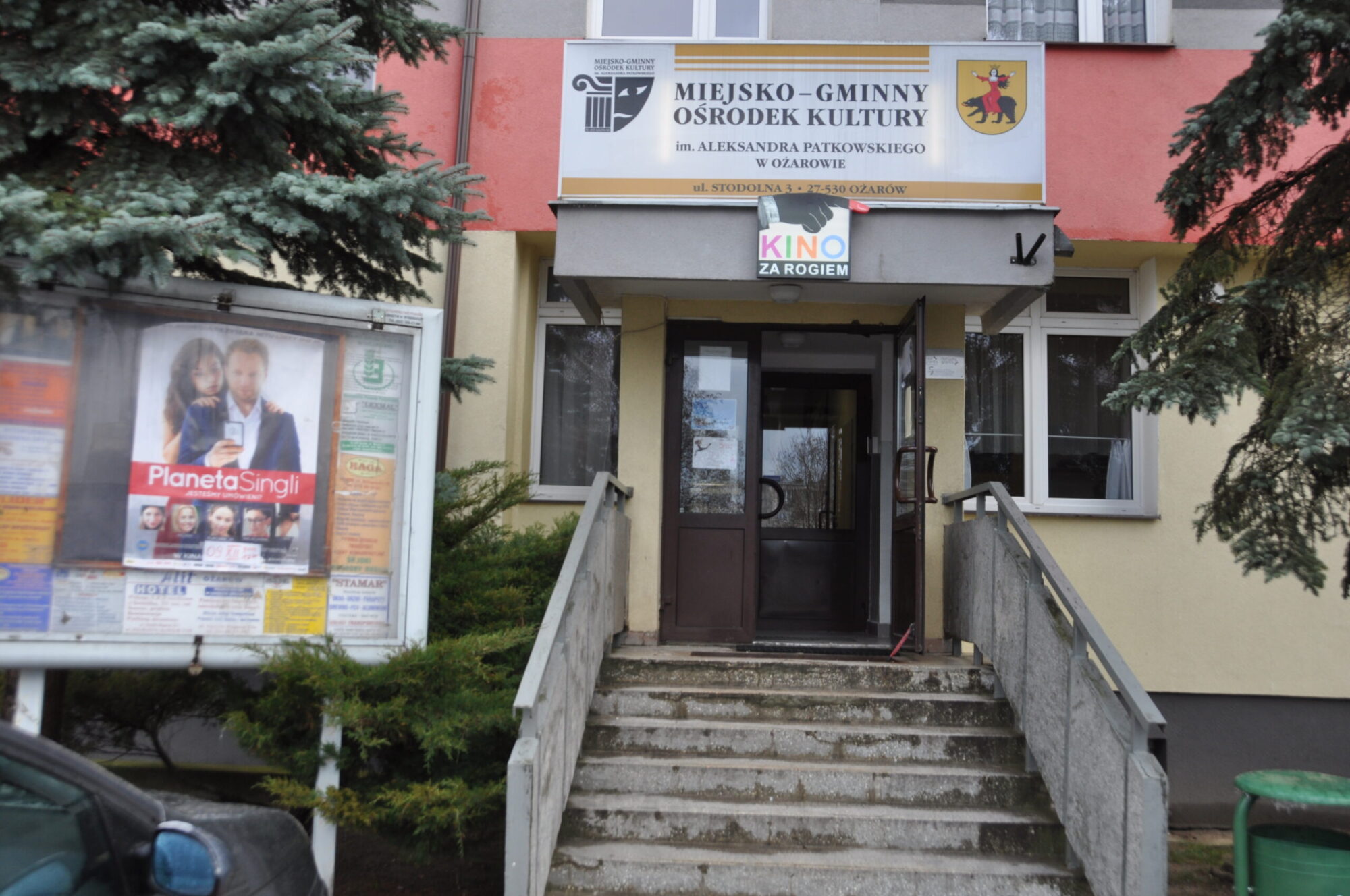 Wejście do centrum kulturalnego w Polsce z rampą i schodami, tablica ogłoszeń z plakatami po lewej stronie i znak nad drzwiami z napisem Kino za Rogiem. Drzewa obramowują scenę.