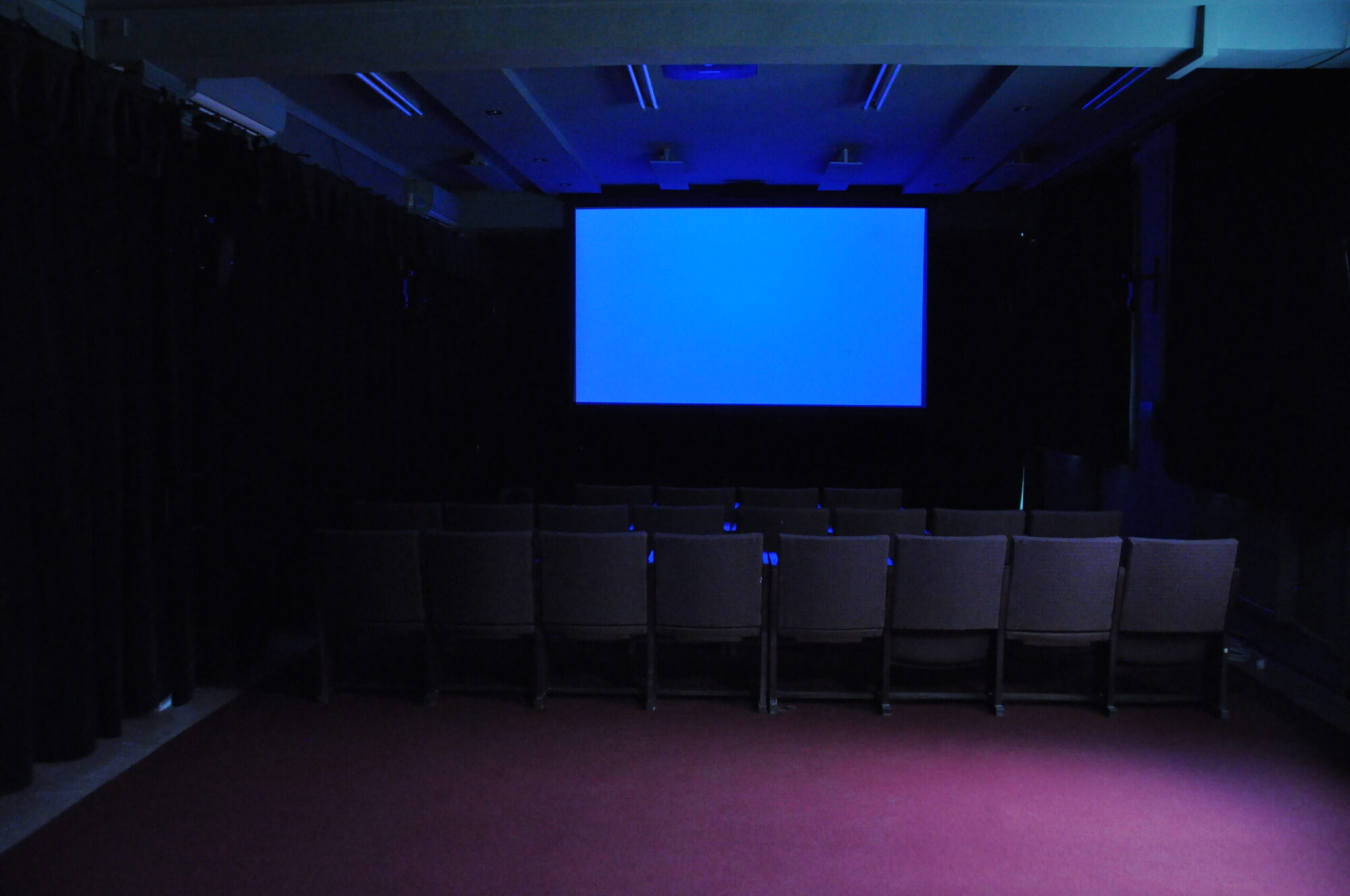 Mały ciemny teatr z pustymi rzędami krzeseł naprzeciwko dużego ekranu wyświetlającego niebieski obraz. Pomieszczenie jest słabo oświetlone, z czarnymi zasłonami po bokach i bordowym dywanem na podłodze.