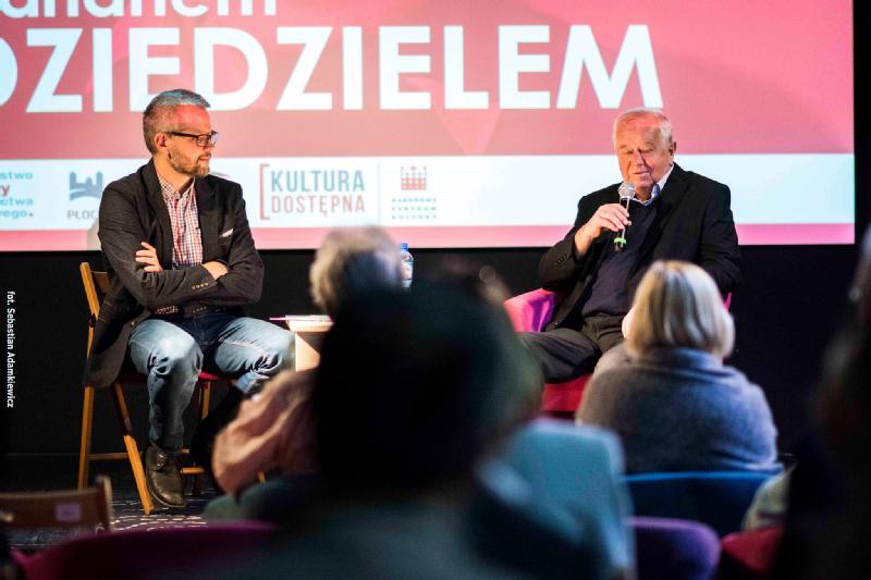 Dwóch mężczyzn siedzi na scenie z mikrofonami, angażując się w dyskusję przed publicznością. Za nimi wyświetla się duży znak z polskim tekstem, a także logo wydarzenia i sponsorów.