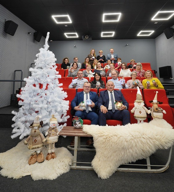 Grupa ludzi pozuje do zdjęcia w małym teatrze z czerwonymi siedzeniami, obok białej choinki udekorowanej ozdobami, w otoczeniu świątecznych pluszowych figurek i sań przykrytych białym futrzanym dywanem.