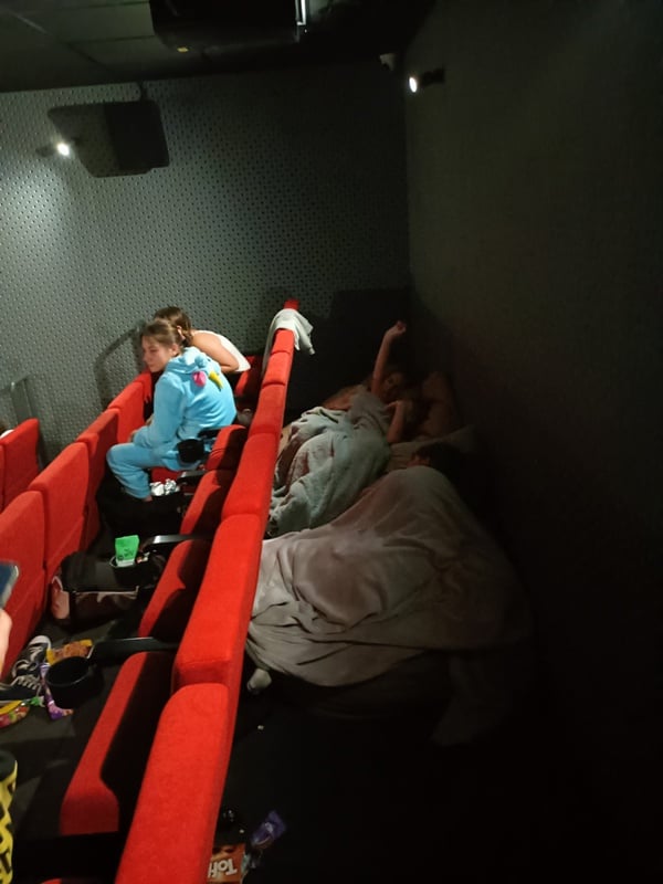 Kilka osób, niektóre owinięte w koce, a jedna w niebieskim kombinezonie, wyleguje się na czerwonych siedzeniach w słabo oświetlonym kinie, co tworzy swobodną i przytulną atmosferę.