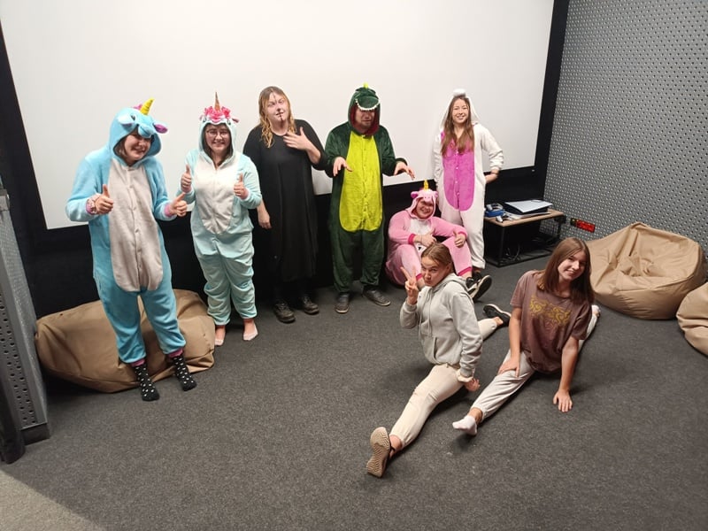 Grupa ośmiu osób pozuje w pokoju, część stoi, część siedzi, wszyscy ubrani są w kolorowe, zwierzęce kombinezony lub codzienne ubrania, na tle wiszą worki sako i duży ekran.