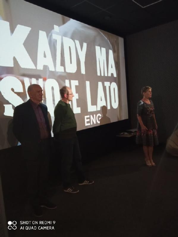 Trzy osoby stoją przed dużym ekranem wyświetlającym polski tekst „KAŻDY MA SWOJE LATO” i „ENG”. Pomieszczenie jest słabo oświetlone, co sugeruje pokaz filmu lub prezentację.