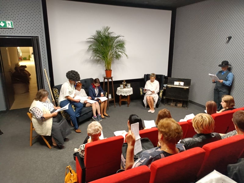 Grupa ludzi siedzi na scenie i czyta scenariusze przed publicznością w małym teatrze. Niektórzy siedzą na kanapie, inni na krzesłach, a w tle widać dużą roślinę. Widzowie trzymają papiery.