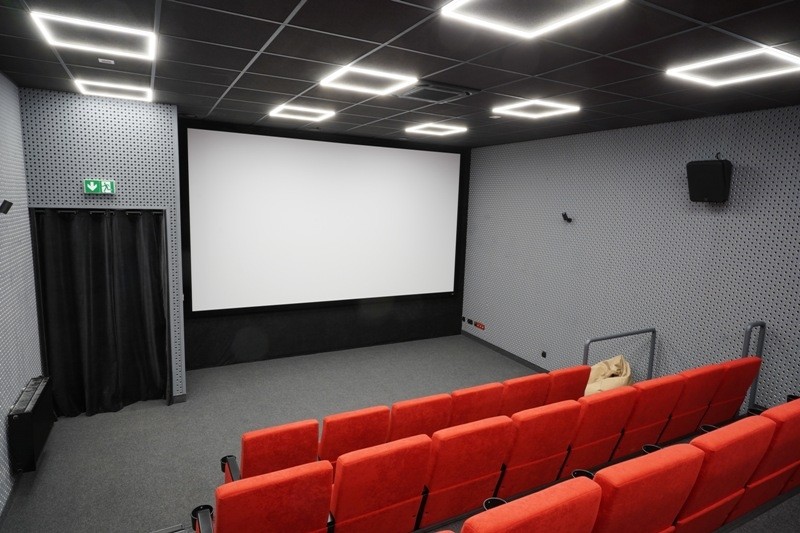 Małe, nowoczesne kino z czerwonymi siedzeniami zwróconymi w stronę dużego, pustego ekranu. Ściany są szare z subtelnym wzorem, sufit ma kwadratowe światła, a po lewej stronie znajdują się drzwi wyjściowe z zielonym znakiem.