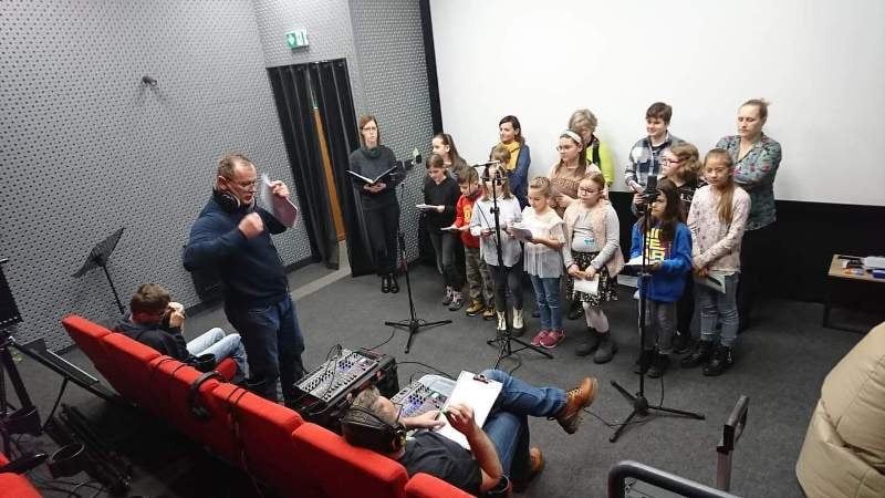 Grupa dzieci i kilku dorosłych stoi przed mikrofonami, ćwicząc w małym studiu nagraniowym, podczas gdy mężczyzna dyryguje grupą, a inna osoba obsługuje sprzęt nagłaśniający w pobliżu czerwonych siedzeń w teatrze.