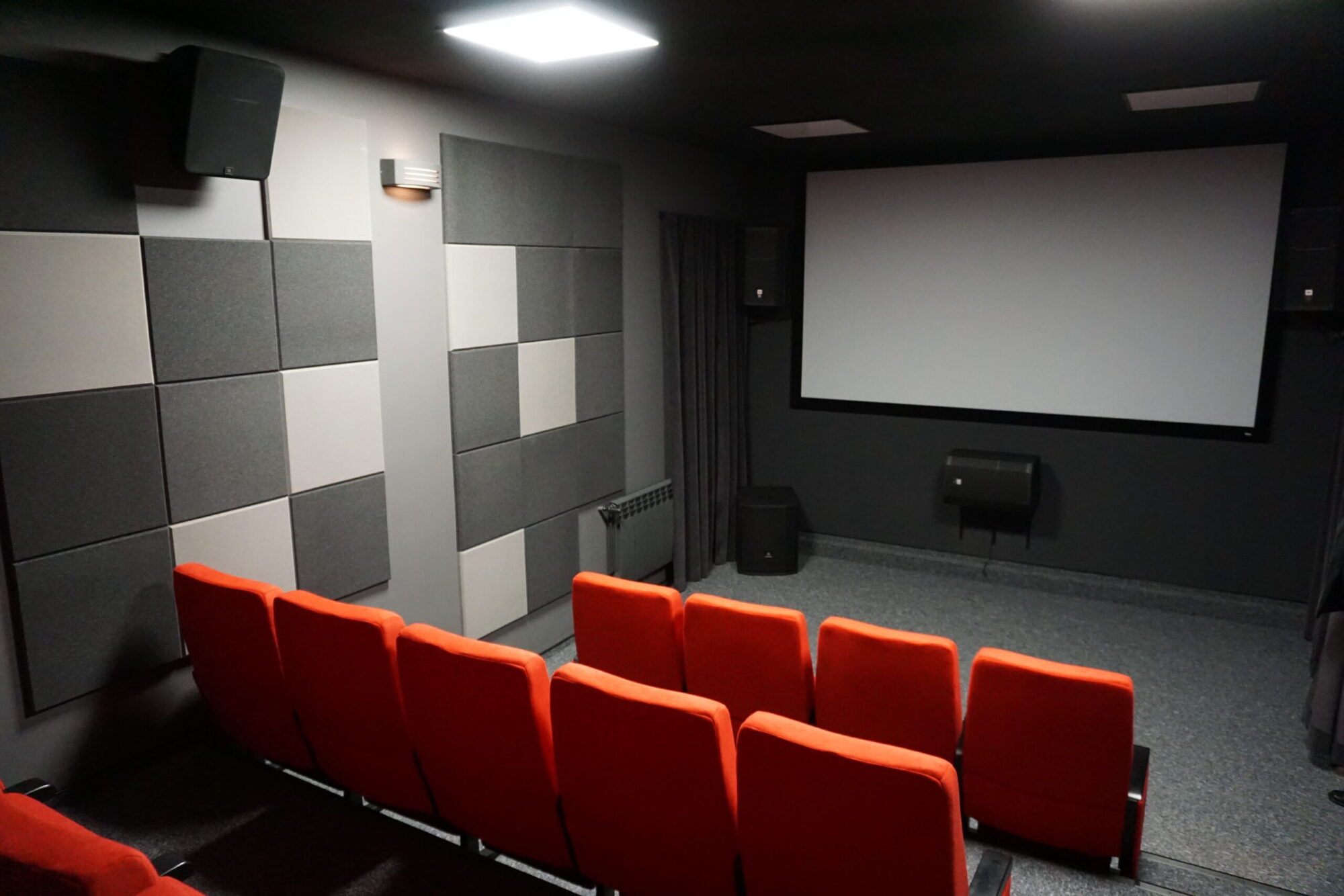 Małe kino z czerwonymi siedzeniami, dużym białym ekranem projekcyjnym, szarymi panelami akustycznymi na ścianach i głośnikami w rogach, widziane z tyłu sali.