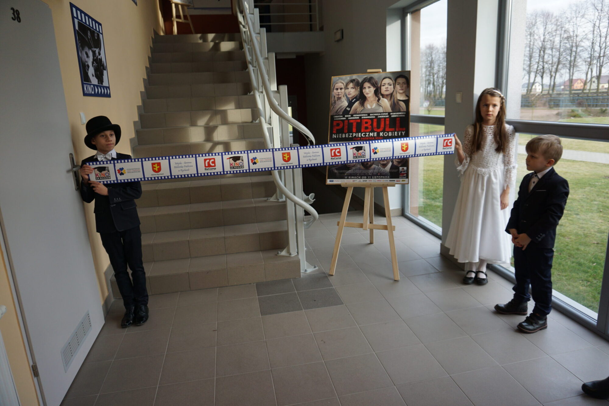 Trójka dzieci ubranych formalnie stoi w pomieszczeniu przy schodach. Dwoje trzyma ozdobną wstążkę z symbolami związanymi z filmem. Za nimi znajduje się plakat filmu Pitbull: Niebezpieczne Kobiety. Duże okna pokazują trawę na zewnątrz.