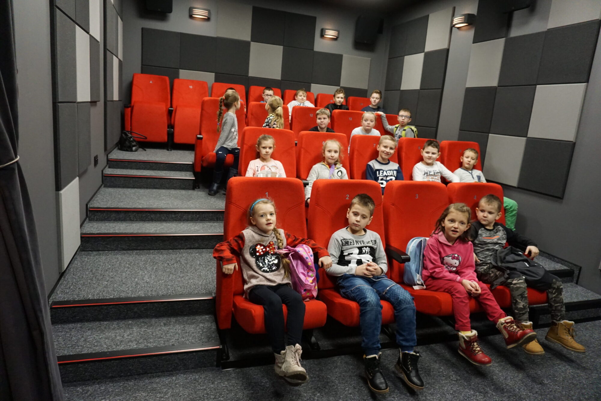 Grupa dzieci siedzi na czerwonych krzesłach w małym kinie lub sali projekcyjnej, zwrócona twarzą do przodu. Ściany są szare z kratkowanym wzorem, a atmosfera jest jasna i zrelaksowana.