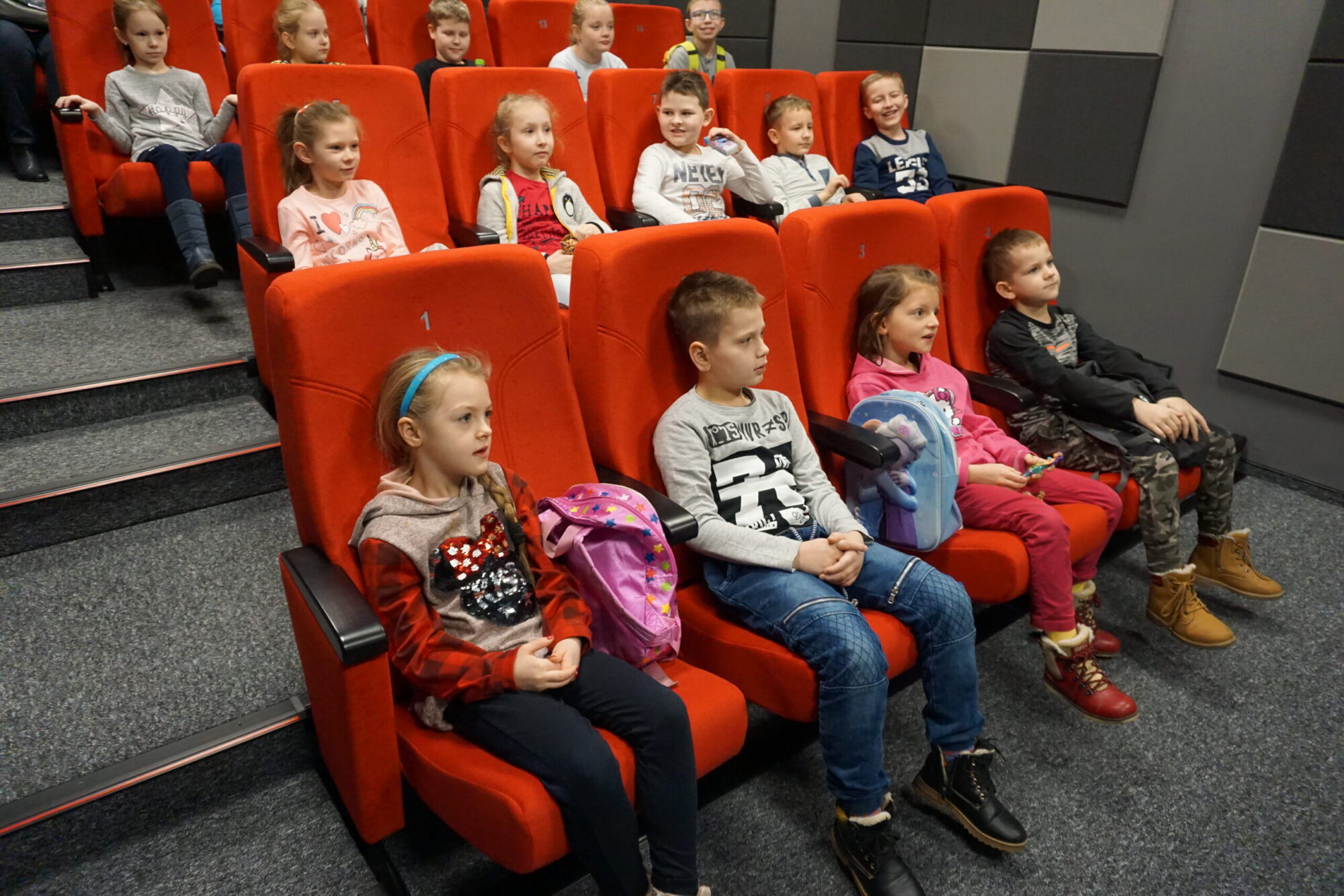 Grupa małych dzieci siedzi na czerwonych fotelach teatralnych, zwrócona twarzą do przodu i uważnie obserwująca. Niektóre mają plecaki na kolanach. Pomieszczenie ma szare ściany z panelami akustycznymi.