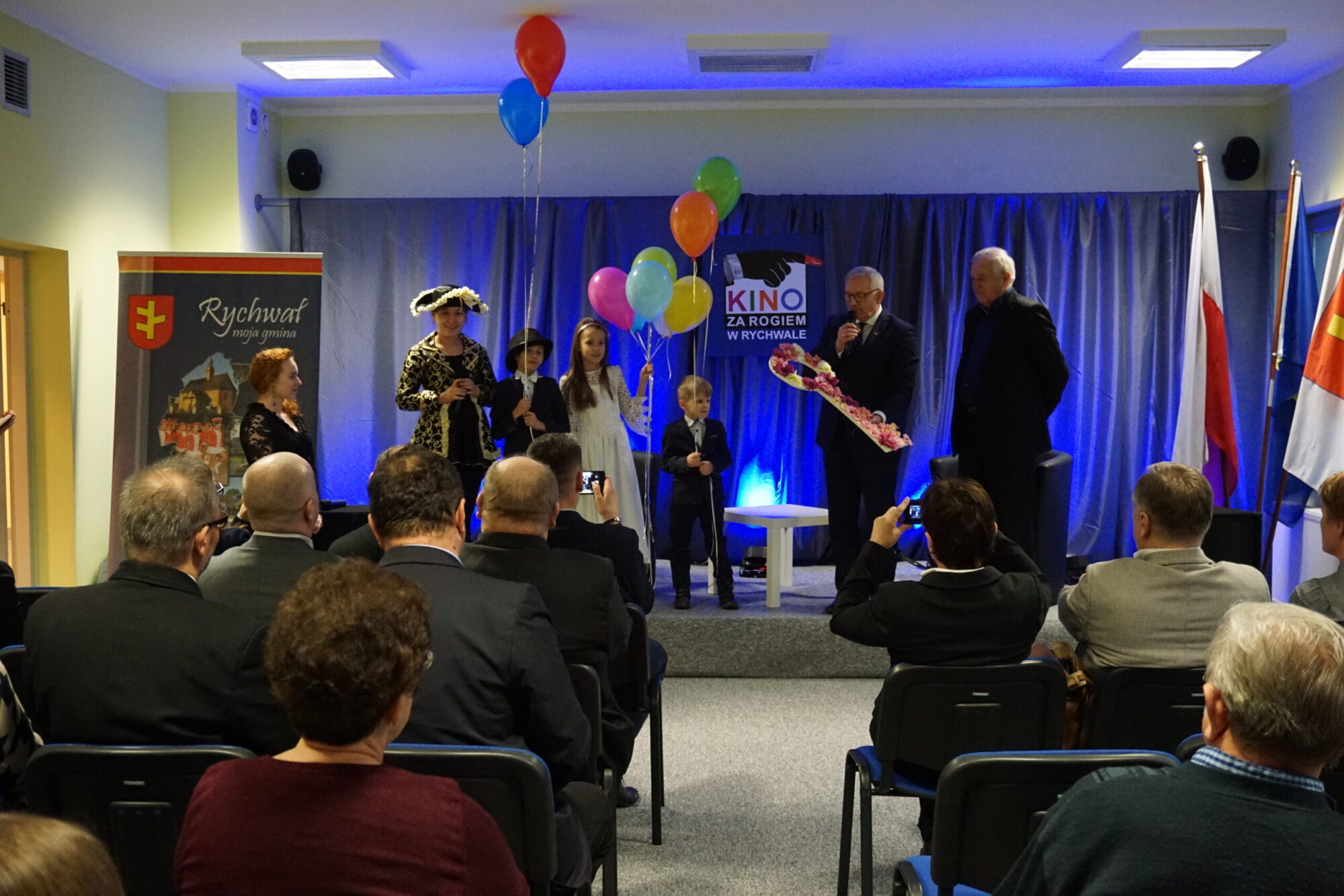 Grupa dzieci trzymających balony i dorośli na scenie są obserwowani przez publiczność w małym audytorium. Jeden mężczyzna trzyma bukiet kwiatów. W tle widoczne są flagi i baner Kino za Rogiem.
