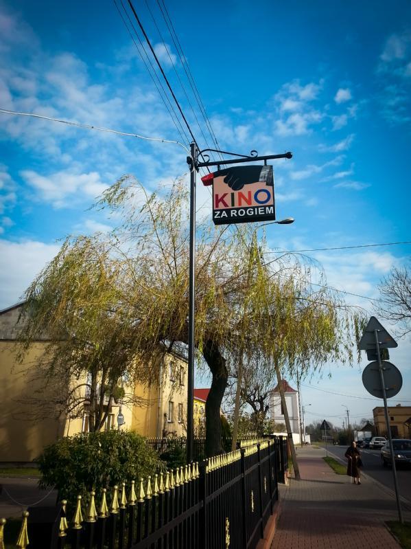 Scena uliczna przedstawia tabliczkę z napisem Kino za rogiem wiszącą na słupie, obok drzewo, żółte budynki, czarny płot i pieszego idącego chodnikiem pod błękitnym niebem z rozproszonymi chmurami.
