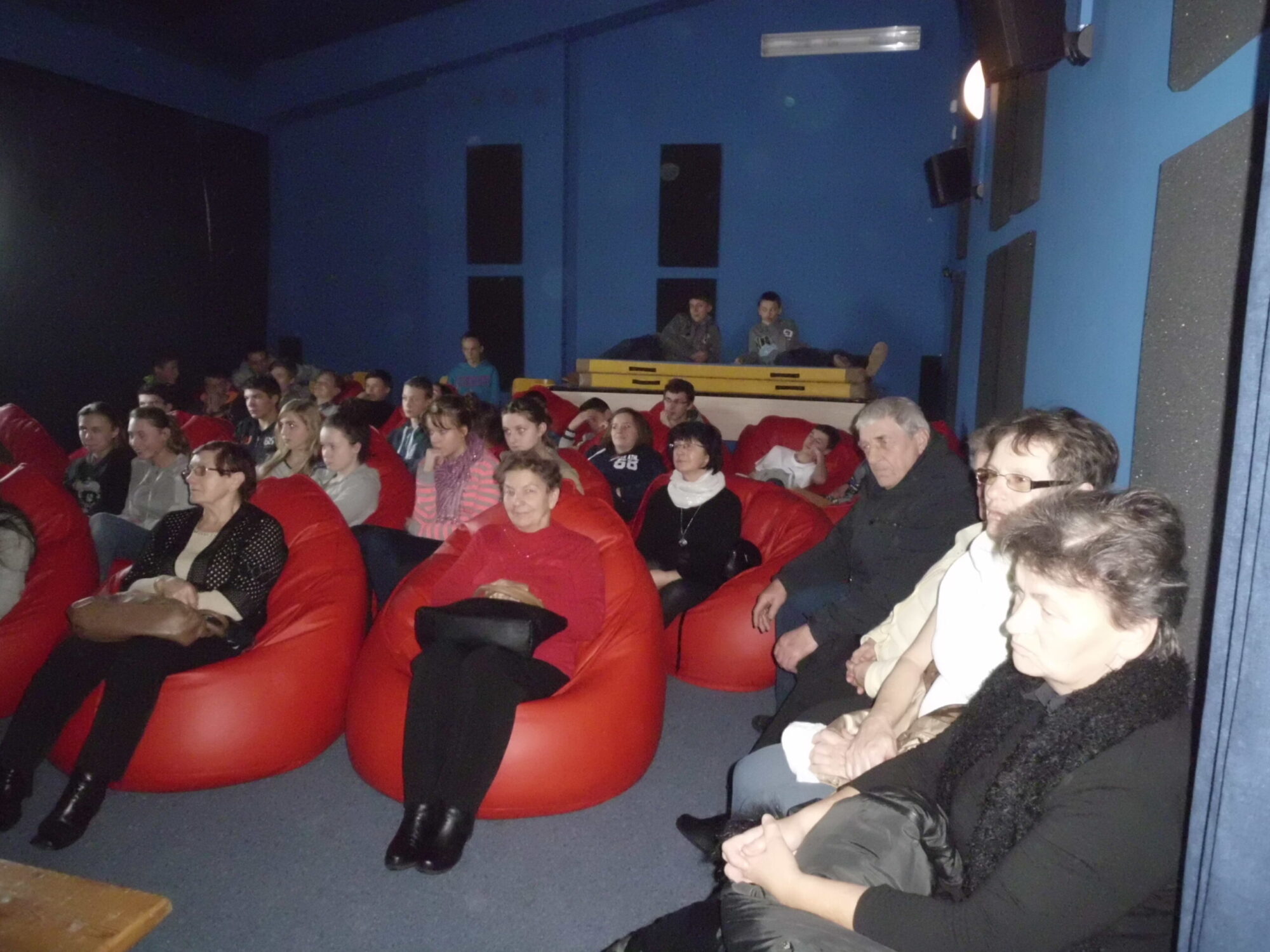 Grupa ludzi siedzi na czerwonych workach z fasolą i krzesłach w słabo oświetlonym pomieszczeniu, uważnie obserwując coś, co nie znajduje się w kadrze. Ściany są niebieskie, a z tyłu siedzi kilka osób.