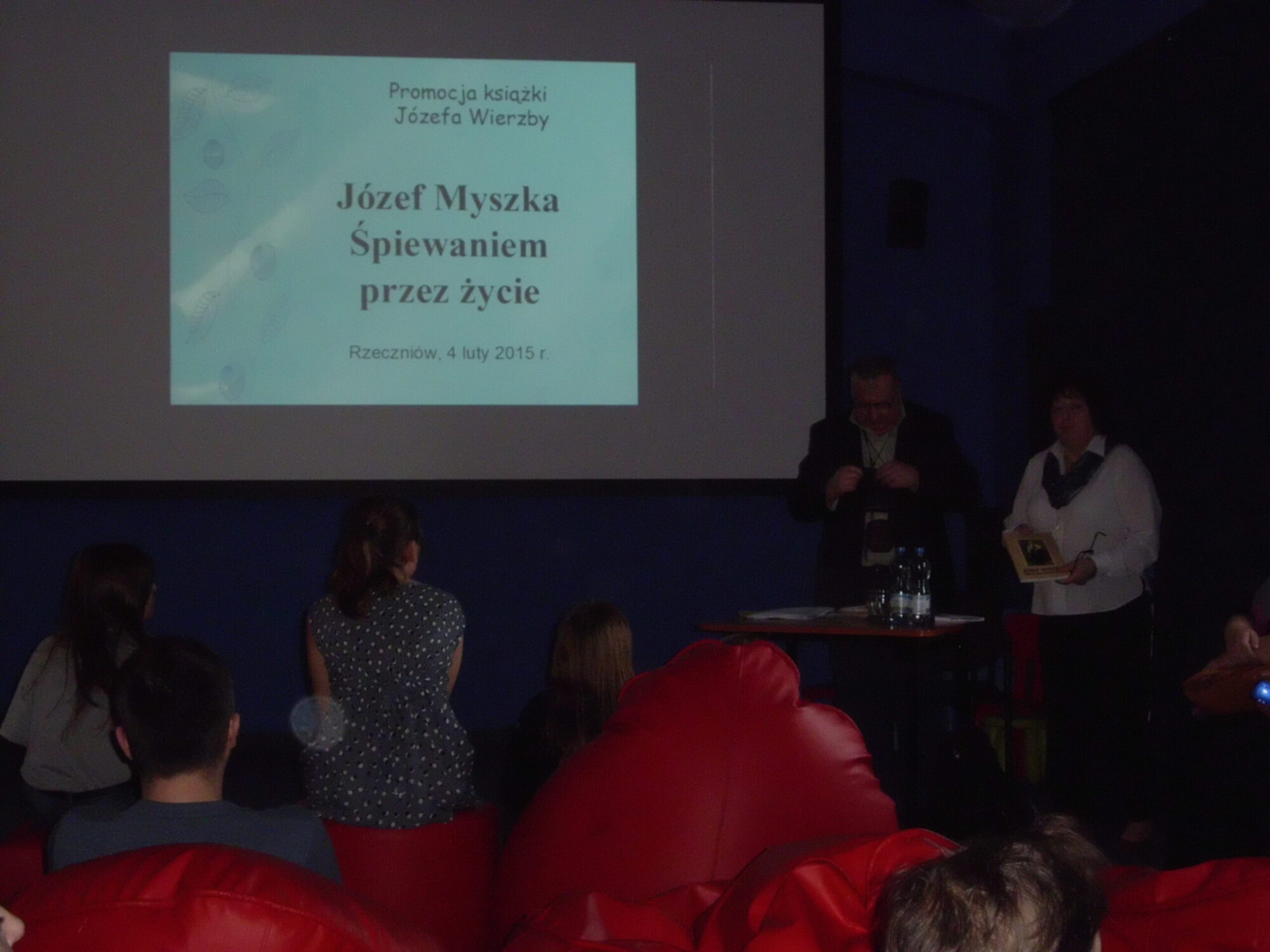 Grupa osób siedzi naprzeciwko ekranu wyświetlającego prezentację zatytułowaną Józef Myszka Śpiewaniem przez życie. Dwóch prezenterów stoi przy stole pod ekranem w słabo oświetlonym pomieszczeniu.