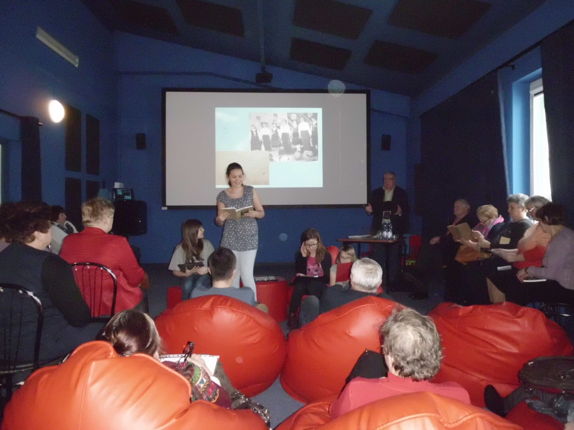 Kobieta stoi i przemawia przed publicznością siedzącą na workach fasolowych i krzesłach w słabo oświetlonym pomieszczeniu. Za nią projektor wyświetla czarno-białe zdjęcie na ekranie, podczas gdy inna osoba stoi w pobliżu.