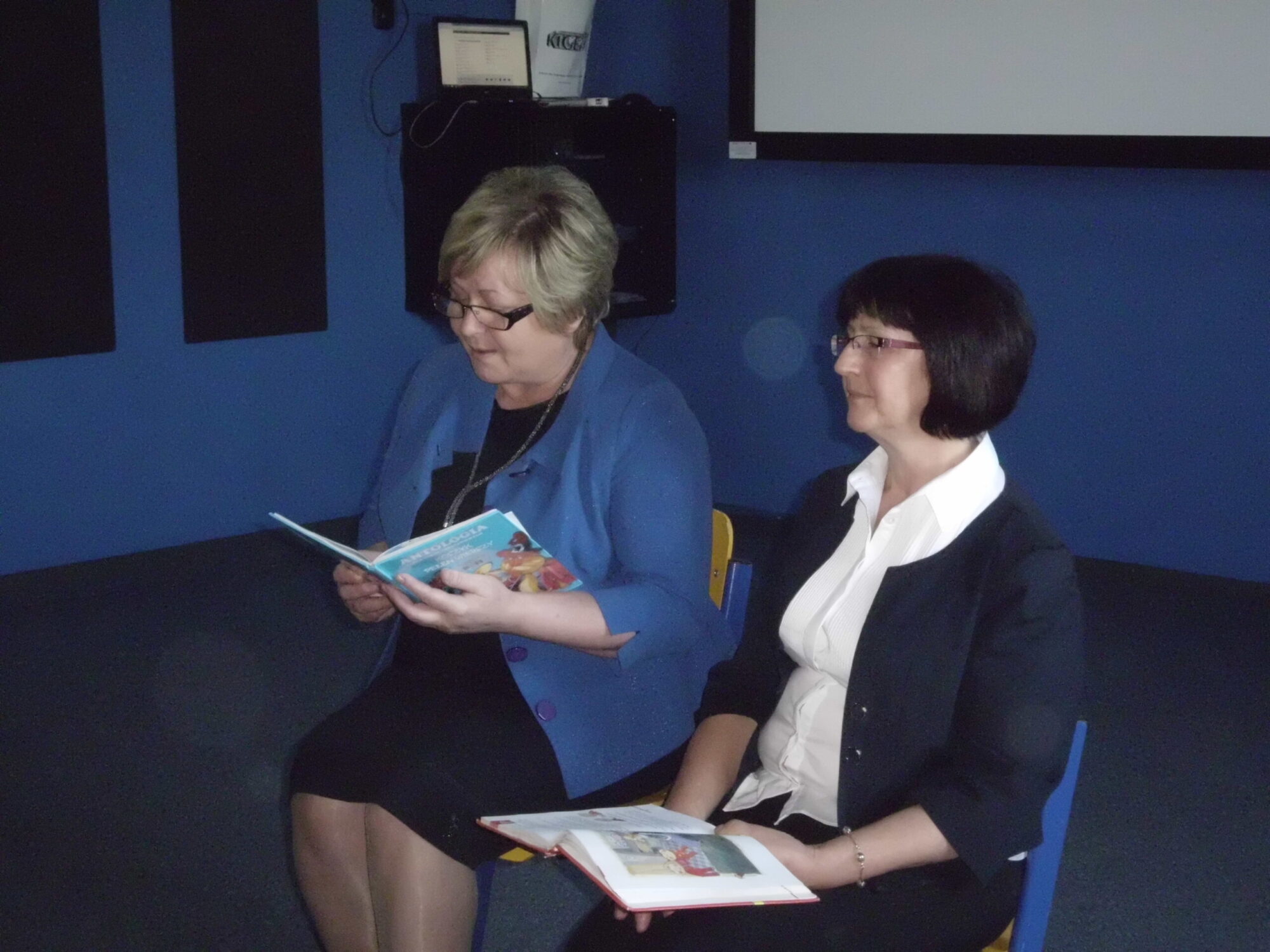 Dwie kobiety siedzą obok siebie na krzesłach w pokoju; jedna czyta otwartą książkę dla dzieci, a druga trzyma książkę na kolanach. Obie są ubrane w stroje biznesowe, a przed nimi znajduje się niebieska ściana i ekran projekcyjny.