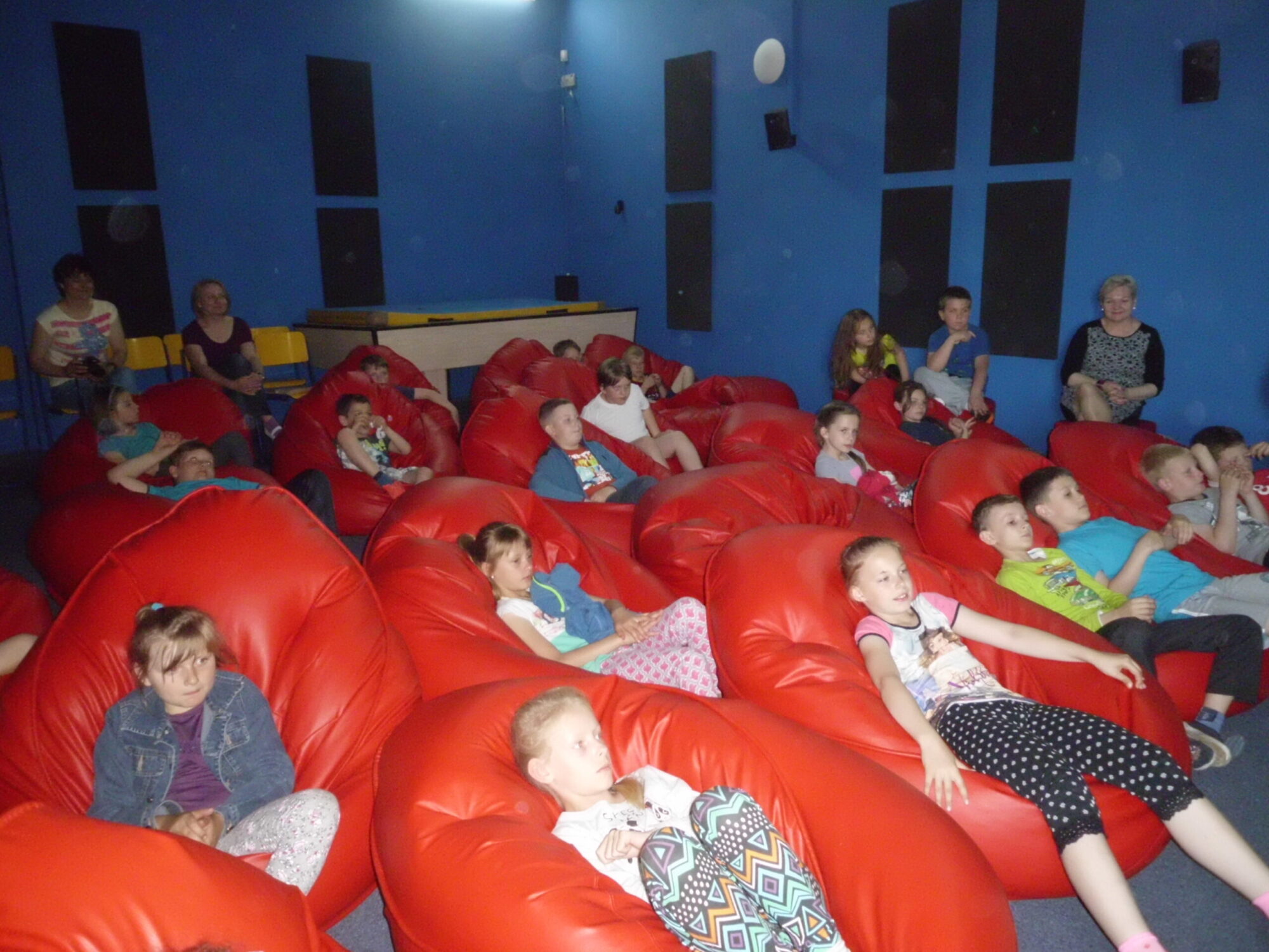 Grupa dzieci siedzi i wyleguje się na dużych czerwonych workach fasoli w pokoju o niebieskich ścianach, uważnie obserwując coś poza kamerą. Kilku dorosłych jest również obecnych w tle.