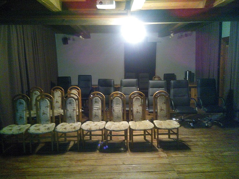 Rzędy pustych krzeseł, w tym wyściełane drewniane krzesła z przodu i krzesła biurowe z tyłu, ustawione w słabo oświetlonym pomieszczeniu z drewnianą podłogą i odsłoniętym sufitem z belkami. Nad głową wisi jasna lampa.