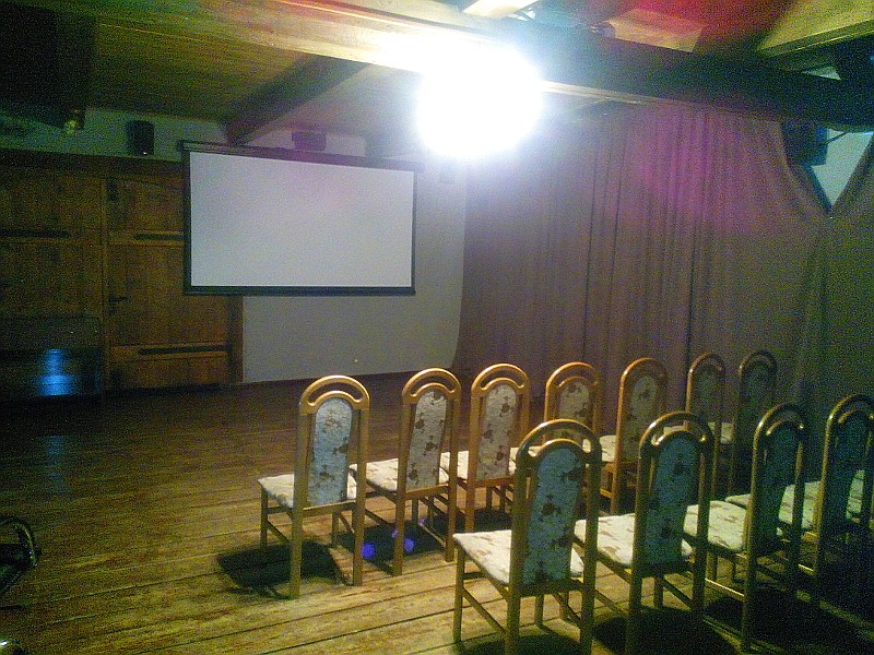 Niewielki pokój z drewnianą podłogą i sufitem, z dwoma rzędami pustych krzeseł ustawionych naprzeciwko pustego ekranu projekcyjnego, oświetlonego jasnym światłem sufitowym.