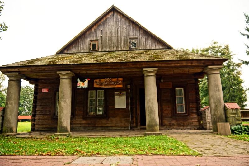 Rustykalny drewniany budynek z trójkątnym dachem, małymi oknami i czterema dużymi kamiennymi kolumnami podtrzymującymi ganek. Nad wejściem widnieje napis Kino, a przed budynkiem jest zieleń i ceglana ścieżka.