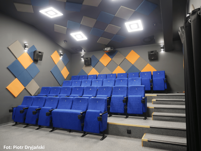 Mała, nowoczesna sala kinowa z niebieskimi, piętrowymi siedzeniami, szarymi ścianami i kolorowymi, dźwiękoszczelnymi panelami w niebieskie i pomarańczowe wzory diamentów. Nad głową znajdują się lampy sufitowe i projektor. Zdjęcie: Piotr Dryjański.
