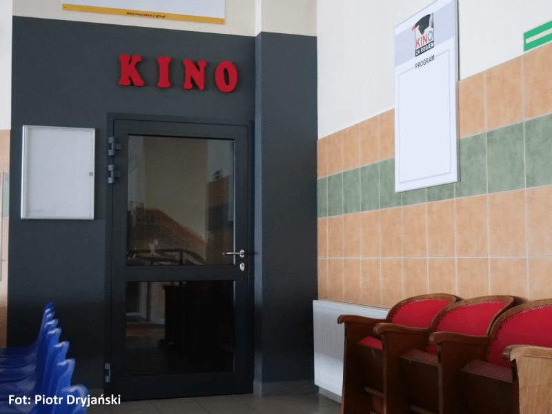 Wejście do małego kina z czerwonymi literami KINO nad czarnymi drzwiami, czerwonymi i niebieskimi krzesłami z boku i kafelkowymi ścianami. Na ścianie wisi biały szyld z logo filmu.