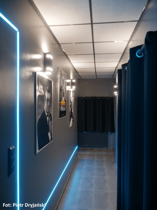 Nowoczesny korytarz z szarymi ścianami, niebieskim oświetleniem LED, oprawionymi plakatami na lewej ścianie i czarnymi zasłonami na końcu. Oświetlenie sufitowe oświetla przestrzeń. Autor zdjęcia: Piotr Dryjański.